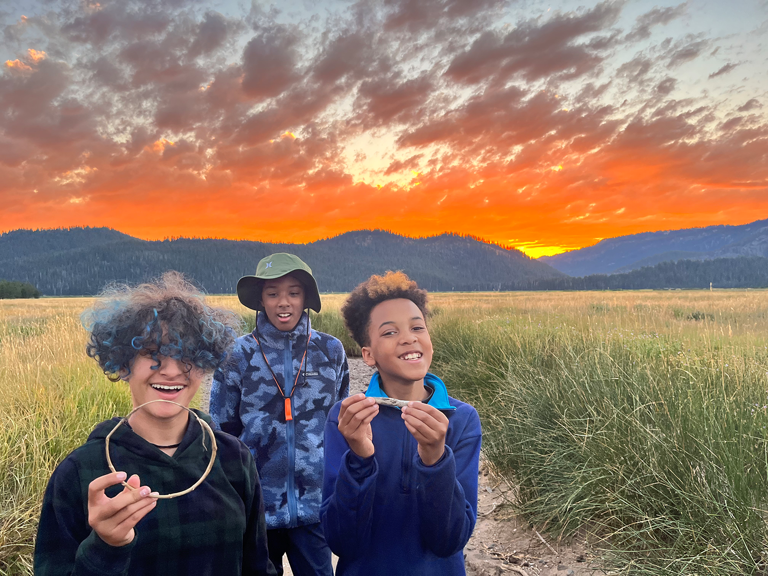 Three young people stand in front of a sunset and smile at the camera.